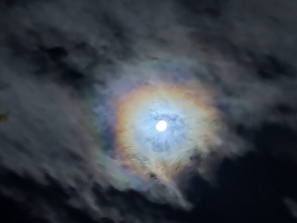 The moon glowing behind soft clouds with a faint rainbow halo in the night sky
